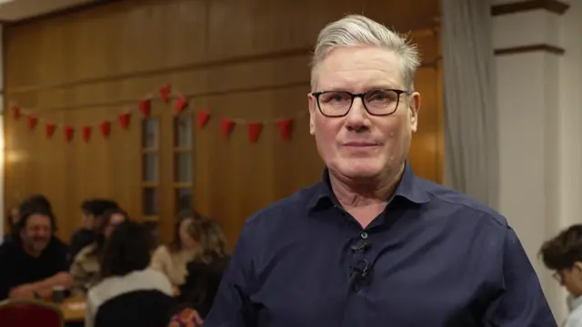 Keir Starmer stands in a hall wearing a blue shirt and looks into the camera