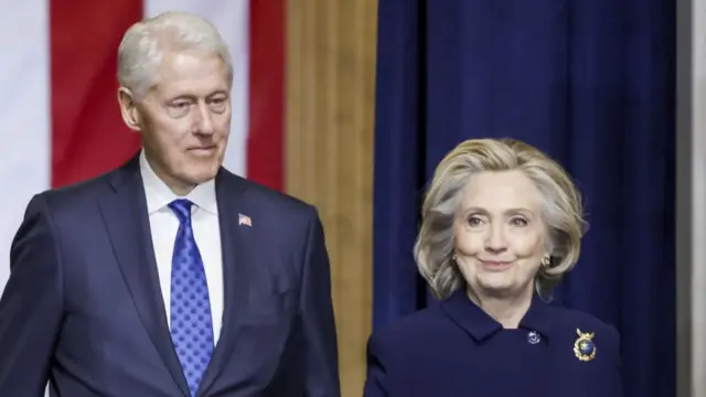 Bill Clinton and Hillary Clinton stand side by side in front of a backdrop of large flags