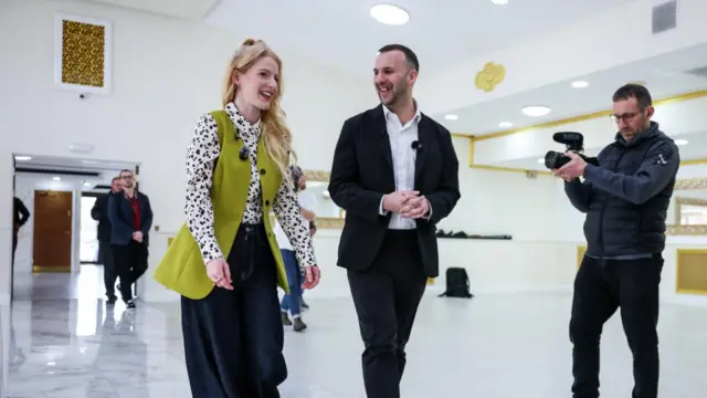 Hannah Spencer wearing a bright green waistcoat and a polka dot shirt walks next to Zack Polanski wearing a dark blazer and a jacket