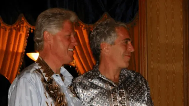 Clinton and Epstein stand next to each other smiling, both wearing shiny shirts
