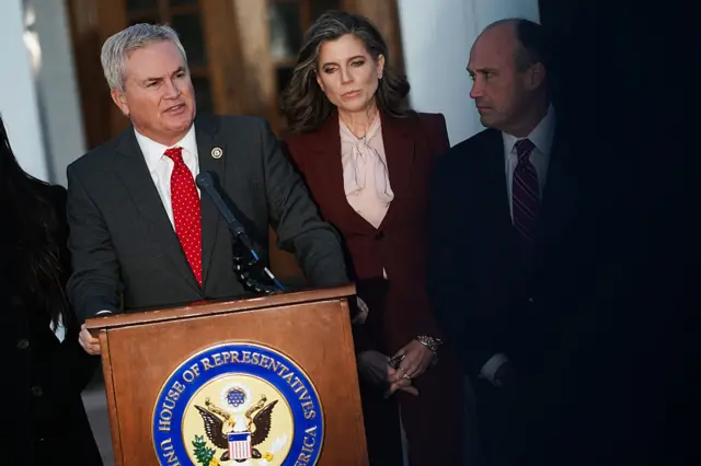 James Comer in a bright red tie stands behind a lectern with Nancy Mace in a burgundy suit standing behind him