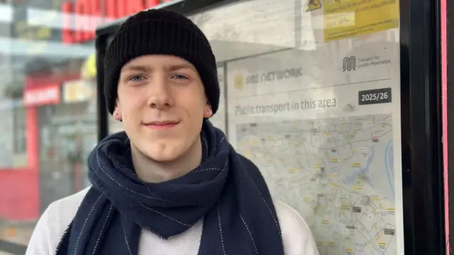Oscar wears a black hat and a blue scarf as he stands at a tram stop in Manchester