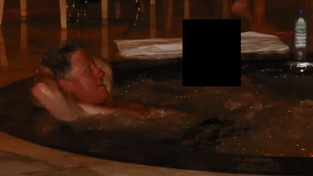 Bill Clinton reclines in a hot tub next to a person whose face has been covered by a black square to protect their identity