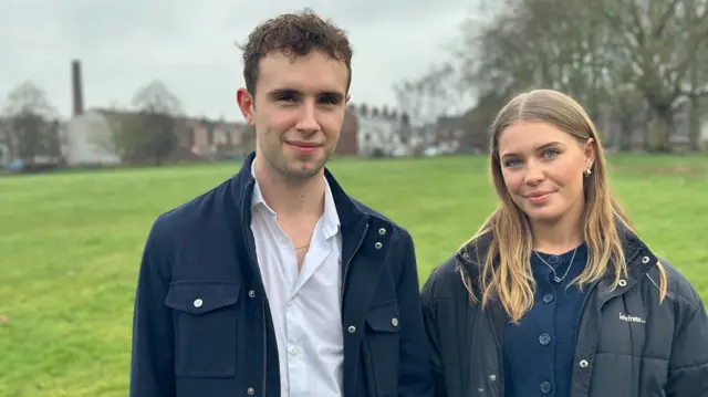 Will wears an open white shirt and a blue jacket and stands next to Rosie who wears a blue shirt and a black puffer jacket
