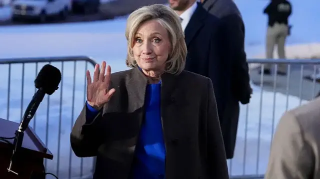 Hillary Clinton, wearing a blue suit and dark coat, smiling and waving in front of a microphone