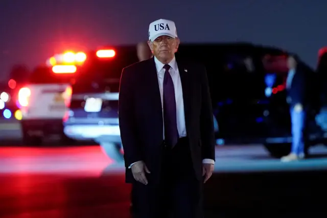 Donald Trump stood on the tarmac at Palm Beach International Airport, there are red car lights behind him and he is wearing a cap that says 'USA'