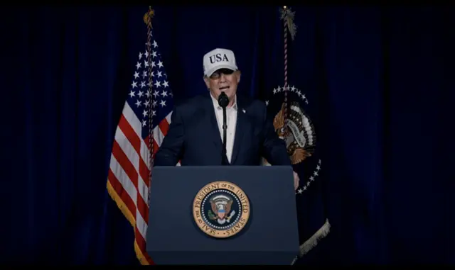 Donald Trump in a suit and white cap marked USA speaks behind a lectern bearing the US President sigil. Behind him a US flag