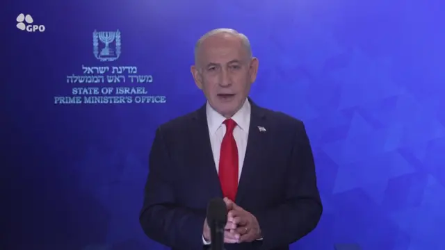 Bebjamin Netanyahu in dark suit, white shirt and red tie speaks into a mic placed in front of him as he stands in front of a blue wall bearing the symbol of Israel and the phrase State of Israel Prime Minister's Office