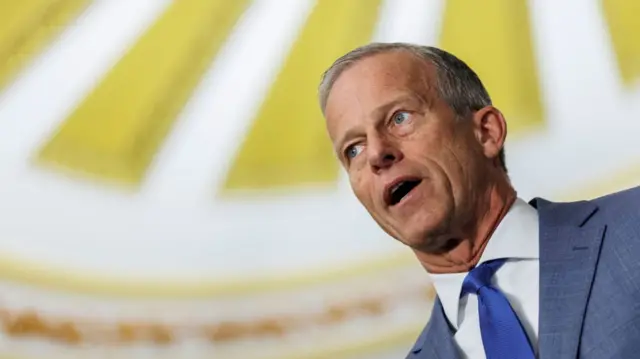 John Thune in light grey suit, white shirt and blue tie mid-speech