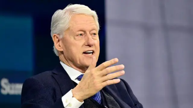 Bill Clinton, wearing a dark blue suit and striped blue tie, gestures with his right hand as he speaks