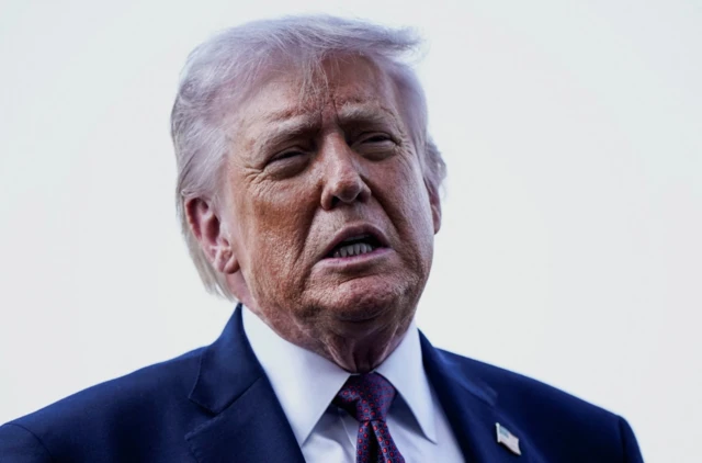 Mid-shot of Donald Trump mid-speech. He's in a dark blue suit, white background behind him
