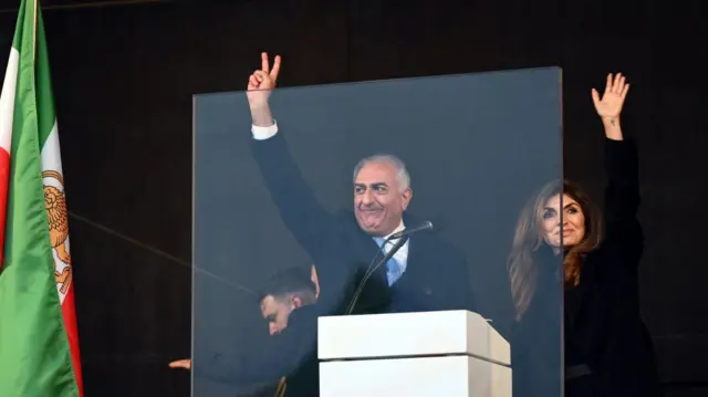 Reza Pahalavi (L) holds up his right arm as he waves at supporters from behind a clear plastic screen. His wife Yasmine Pahalvi (R) stands next to him also waving