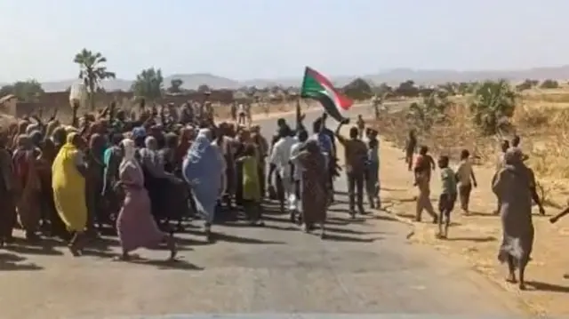 Dozens of people can be seen celebrating on a road - one is carrying the Sudanese flag