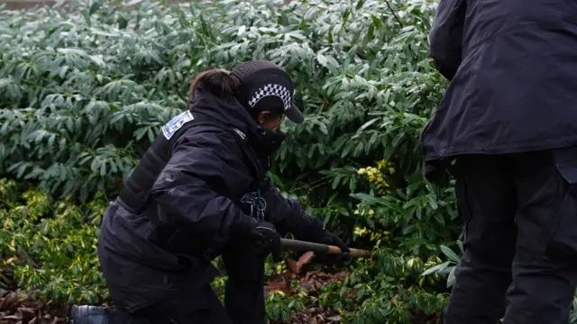 A woman dressed in black police uniform using a stick to search bushes