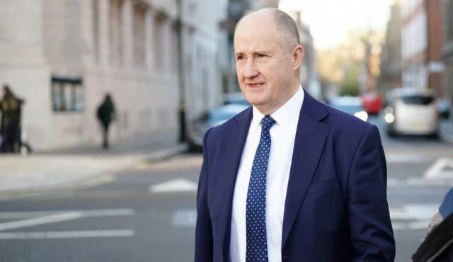 Kevin Hollinrake walks in a central London street. He is wearing a navy suit and tie with a white shirt and is looking away from the camera and in the direction he is walking.