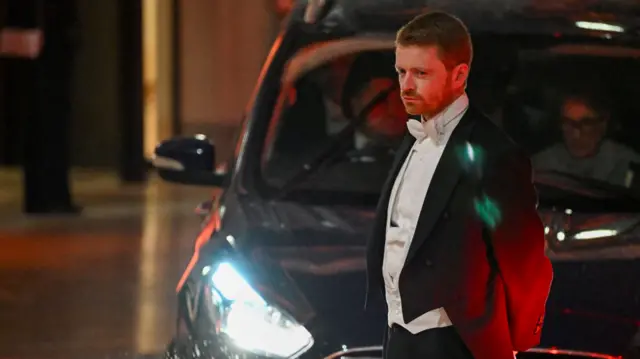 Chief of staff Morgan McSweeney in white tie standing in front of a car with its headlights on and looking moodily into the corner of the frame