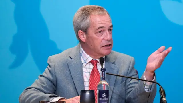 Farage sitting and speaking while sitting one hand on a desk and another raised up. There's a small desk microphone and a glass bottle of water in front of him next to a cup. There's a blue background behind him
