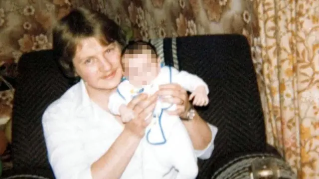 Steve Wright as a younger adult sits in an armchair. He is holding a baby whose face has been blurred. Wright has brown hair and wears a white shirt.