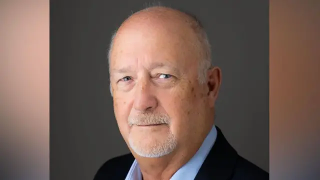 Simon Dinsdale in a portrait-style photo. He angled slightly away from the camera but has turned his head to face the camera and smile. He wears a dark navy suit with a light blue shirt underneath. He is largely bald and has some grey facial hair.