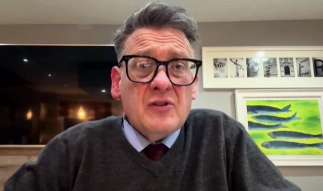 Karl Turner wearing black glasses and a grey jumper with a shirt and tie