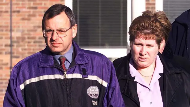 Graham Hall is wearing a shirt and tie with a purple, blue and white anorak over it. He has parted hair and square-shaped glasses. He is walking alongside Lorinda Hall, who wears a pink blouse and neck chain under a black jacket. She has backcombed hair. Behind them is a brick court building.