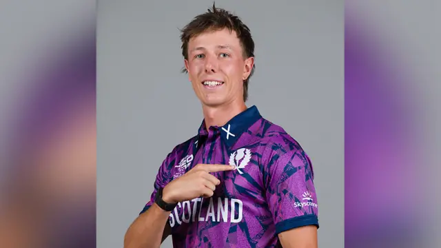 Scotland cricketer Olly Davidson looks at the camera with a finger from his right hand pointing to the white badge on the chest area of his purple and blue patterned Scotland cricketing jersey.