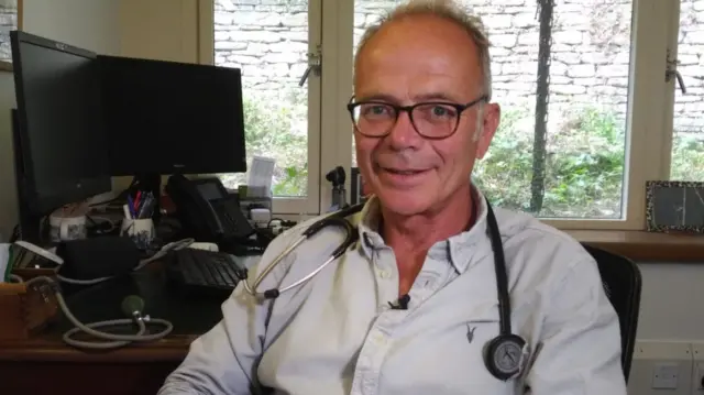 Simon Opher MP sits at his desk with a stethoscope around his neck