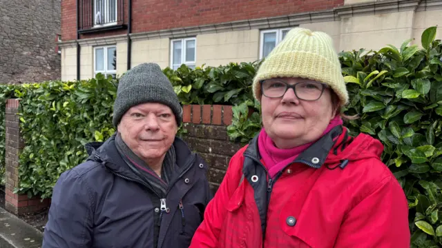 An elderly man wearing a dark coat and hat stands next to a elderly woman with glasses, a red coat and green hat. They are standing on the street in front of a hedge.