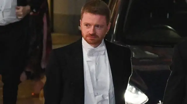 Morgan McSweeney in white tie with his hands behind his back and standing in front of a car with its headlights on