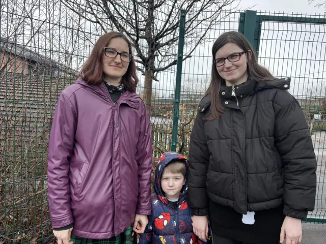 A mother stood outdoors with her daughter and son. They are looking at the camera and smiling.