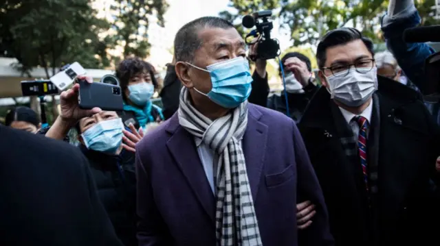 Jimmy Lai wearing a scarf and face mask, surrounded by reporters