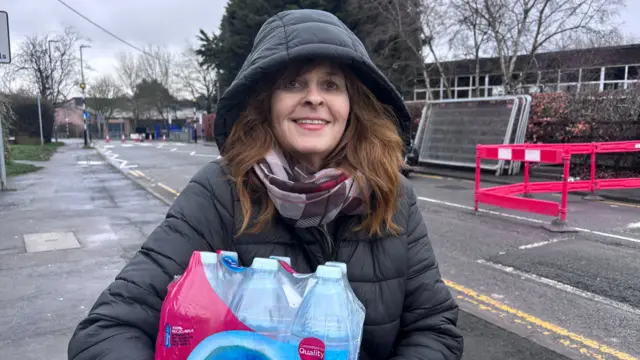 a woman carrying a pack of bottled water. She is wearing a black coat with the hood up and a scarf. She is looking at the camera and smiling.