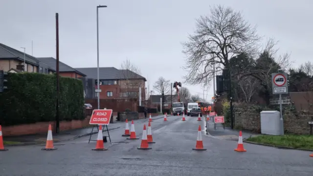 A closed road in Kingswood. There are cones and a road closed sign at the junction. Vehicles and water crews can be seen in the distance.