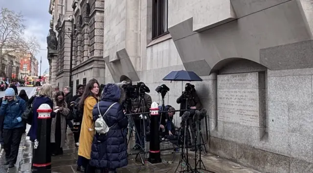 Journalists gathered outside a court on a grey day. Some have cameras and others are holding microphones and mobile phones.