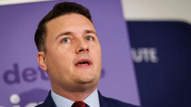 Close up of Health Secretary Wes Streeting mid speech in front of two signs, one in lilac and one in dark blue, blurred behind him