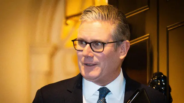 Keir Starmer half smiling as he steps out of Number 10, a folder in his arms. He's in a black suit, white tie and patterned tie, the black front door of the building open behind him showing a glimpse of the residence