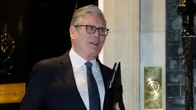 Prime Minister Keir Starmer in a black suit, white shirt and patterned tie walks out of Number 10 holding a black folder in his left hand