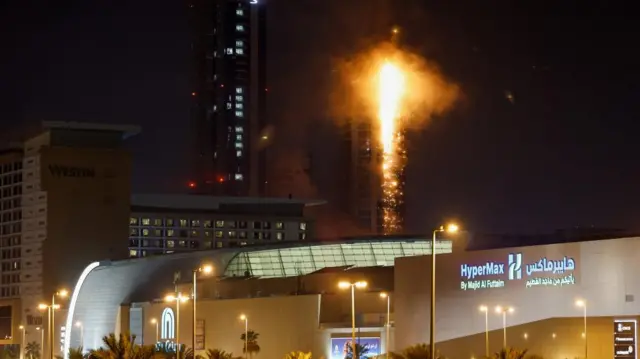A streak of flames runs up the side of a tower block at night. Several buildings can be seen in front of the burning tower
