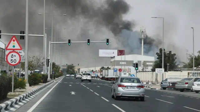 A cloud of grey and black smoke rises can be seen from the side of a highway with two lanes. A silver car is driving near some traffic lights in the front of the picture