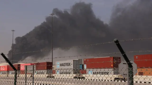 Thick black smoke billows in the backdrop.Red and grey containers are stacked in front of it and there is barbed wire in the front of the photo