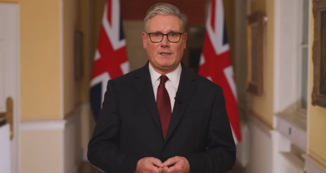 Prime Minister Keir Starmer in a dark suit stands in front of two UK flags