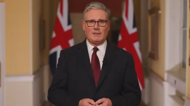 Prime Minister Keir Starmer in a dark suit stands in front of two UK flags