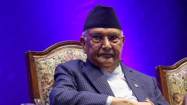 An elderly man with a traditional black hat and wearing a blue checked suit sits in a chair in front of a purple backdrop