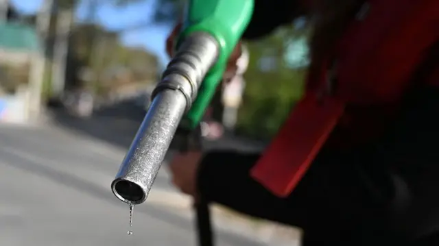 An attendant holds a green fuel pump at a petrol station. A drop of petrol is sliding off the nozzle.