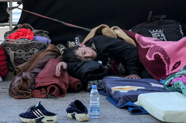 A young boy sleeps on a mattress outdoors with his family's belongings close by.