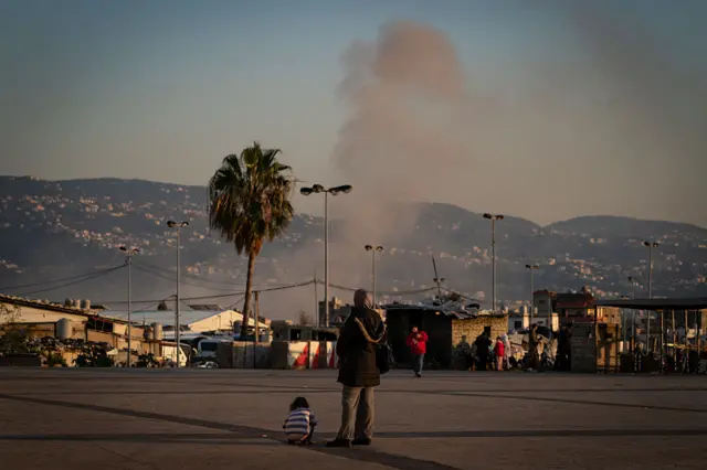 A child sits on the floor at the Camille Chamoun Sports City Stadium. An adult looks on at the smoke rising from a strike in Beirut's southern suburbs