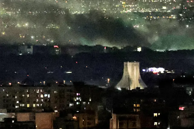 A night-time image of a Tehran cityscape, with smoke rising beyond a lit up Azadi Tower in the right, middle-ground.