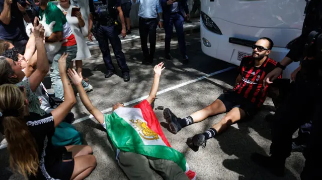 A protester draped in the Iranian flag before the Islamic revolution lays in front of a bus on the pavement in Australia in support of the Iranian women's football team