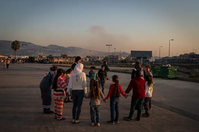 Children hold hands as they play at a shelter on the Camille Chamoun Sports City Stadium in Beirut, Lebanon, as smoke rises from a nearby strike