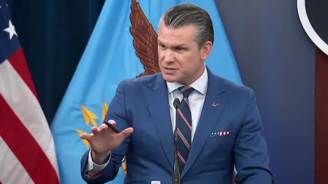 Pete Hegseth in a blue suit, with his right hand raised with a pen in between his fingers, speaking into a microphone on a podium